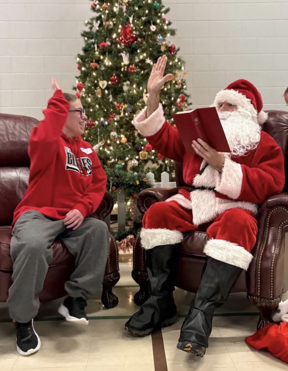 Santa reading while high fiving a member by the Christmas tree.